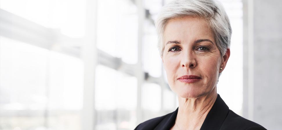 A white-haired, business woman stands near bright office windows.