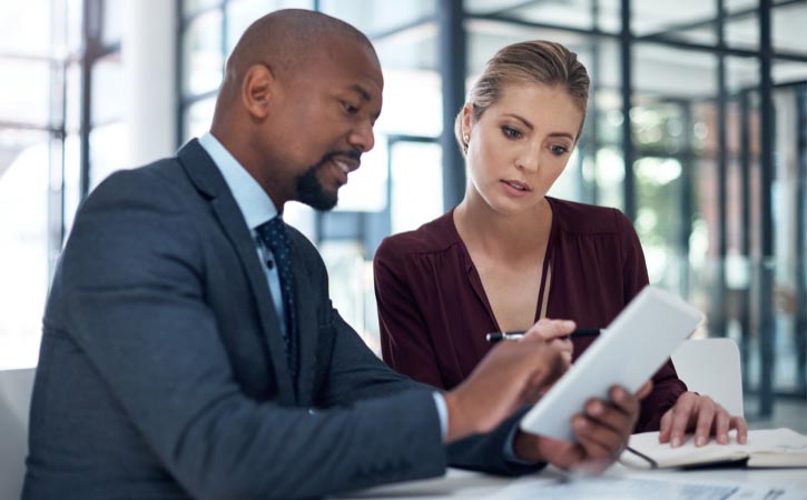 iStock-1183043221-1 Un homme et une femme d’affaires regardent une tablette numérique dans un bureau d’allure moderne.