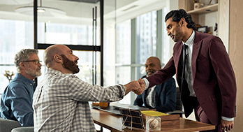 A man in a burgundy suit shakes hands with a seated, bearded man in a plaid shirt at a conference table. Two others men observing.