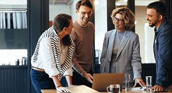Four people stand around a table with a laptop, papers, and drinks, engaged in a discussion and smiling in a modern office setting