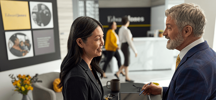 A man and woman in business attire converse and smile in a modern Edward Jones office. Two people walk past a reception desk in the background.