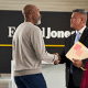 An office setting where two men shake hands, one in a suit holding documents, standing in front of a Edward Jones sign.