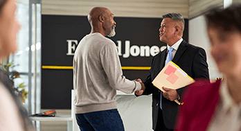 An office setting where two men shake hands, one in a suit holding documents, standing in front of a Edward Jones sign.