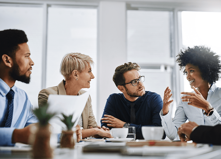 A diverse group of professionals engaged in a lively discussion around a conference table. Bright lighting, plants, and papers create a collaborative atmosphere.