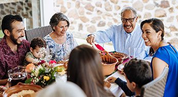 multi-generational family shares a meal outdoors. An older man with glasses serves food from a pot, surrounded by smiling relatives and flowers.