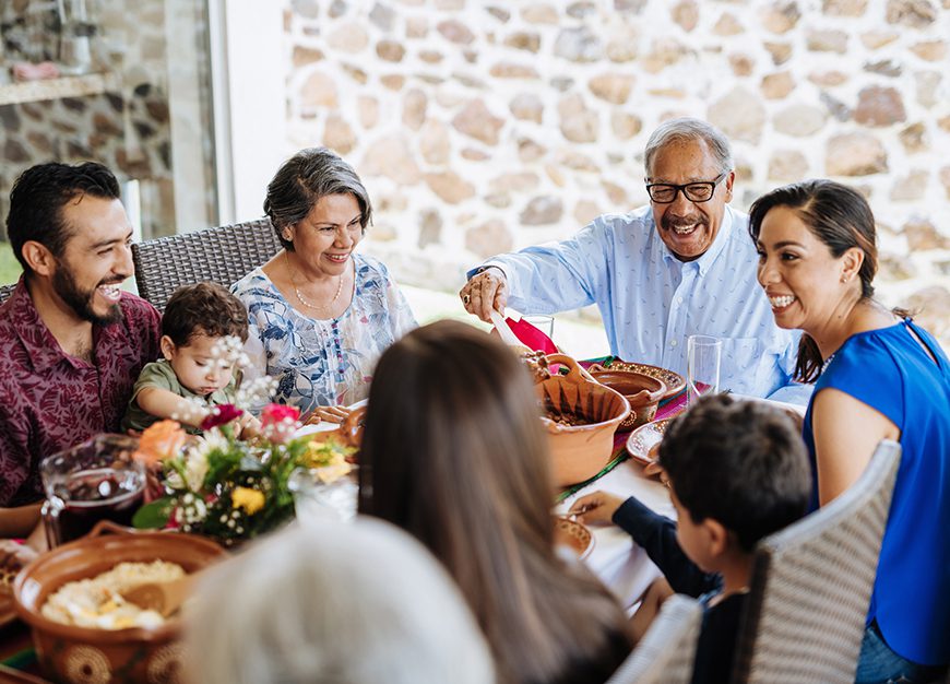 multi-generational family shares a meal outdoors. An older man with glasses serves food from a pot, surrounded by smiling relatives and flowers.