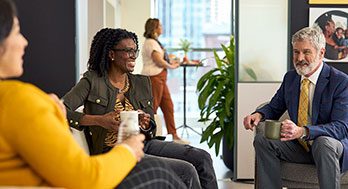 Three people sit and talk in an office lounge, each holding a mug, while another person stands by a counter in the background.