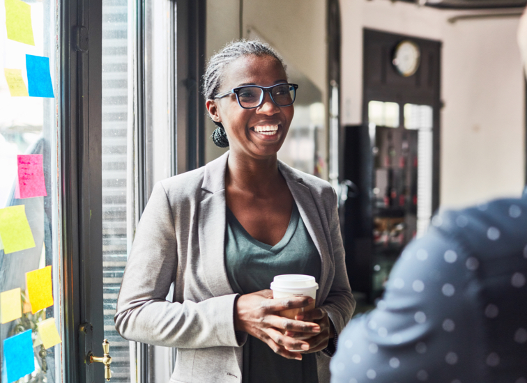 A woman stands near a bright window holds a beverage while speaking with another person.