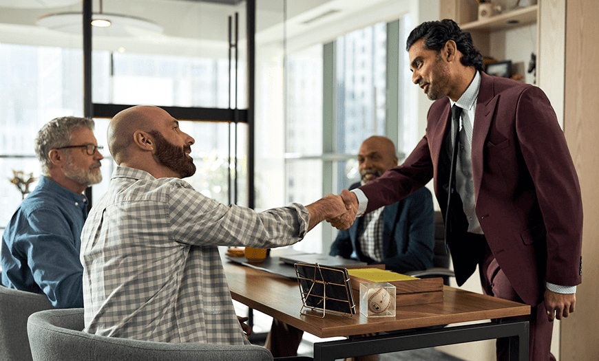 A man in a burgundy suit shakes hands with a seated, bearded man in a plaid shirt at a conference table. Two others men observing.