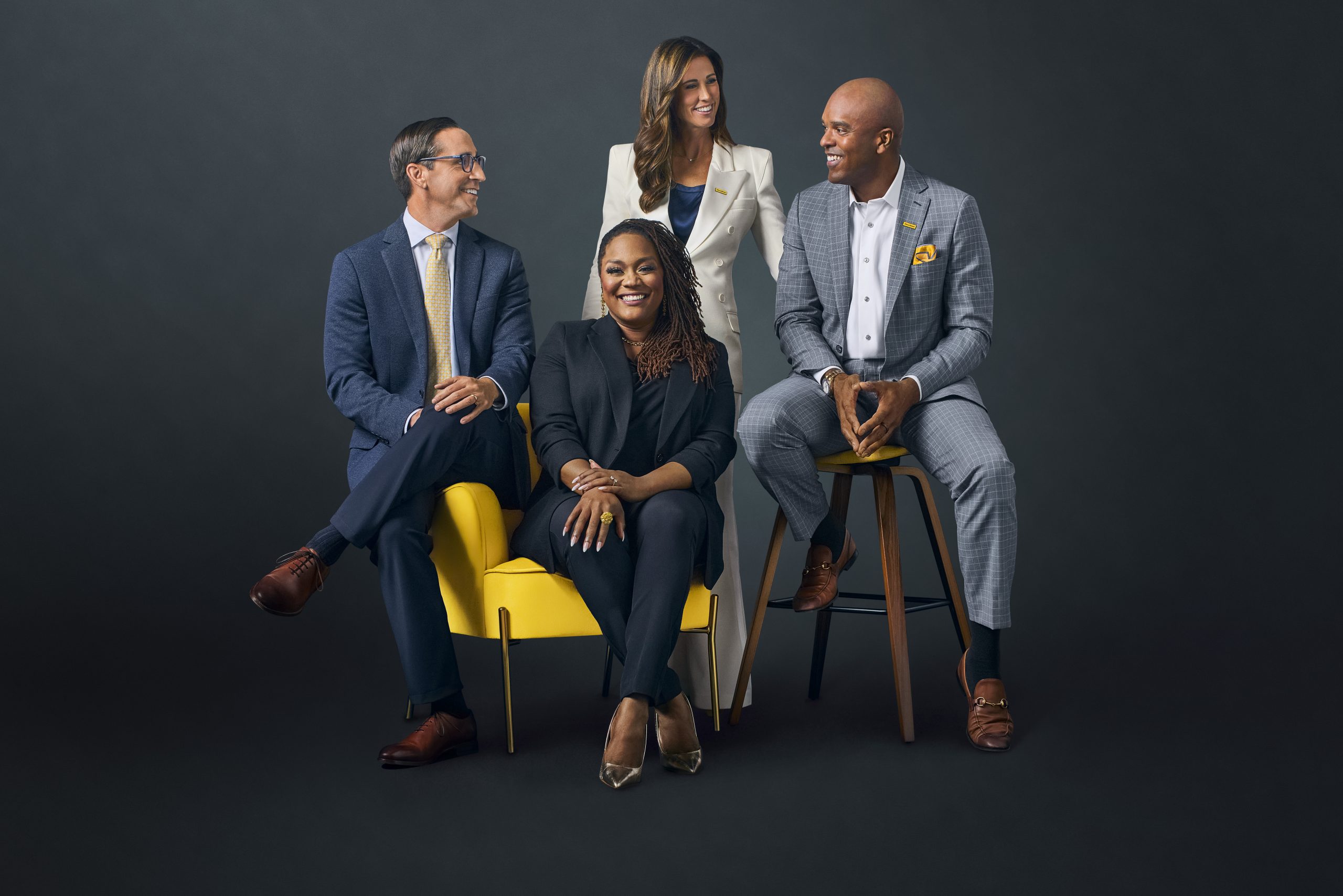 Four professionally dressed colleagues—two seated on a yellow chair, one standing and another seated on a tall wooden stool—smiling at one another.