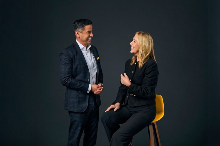 Two professionally dressed colleagues smiling at each other— the man stands and the woman is seated on a yellow stool against a dark background.